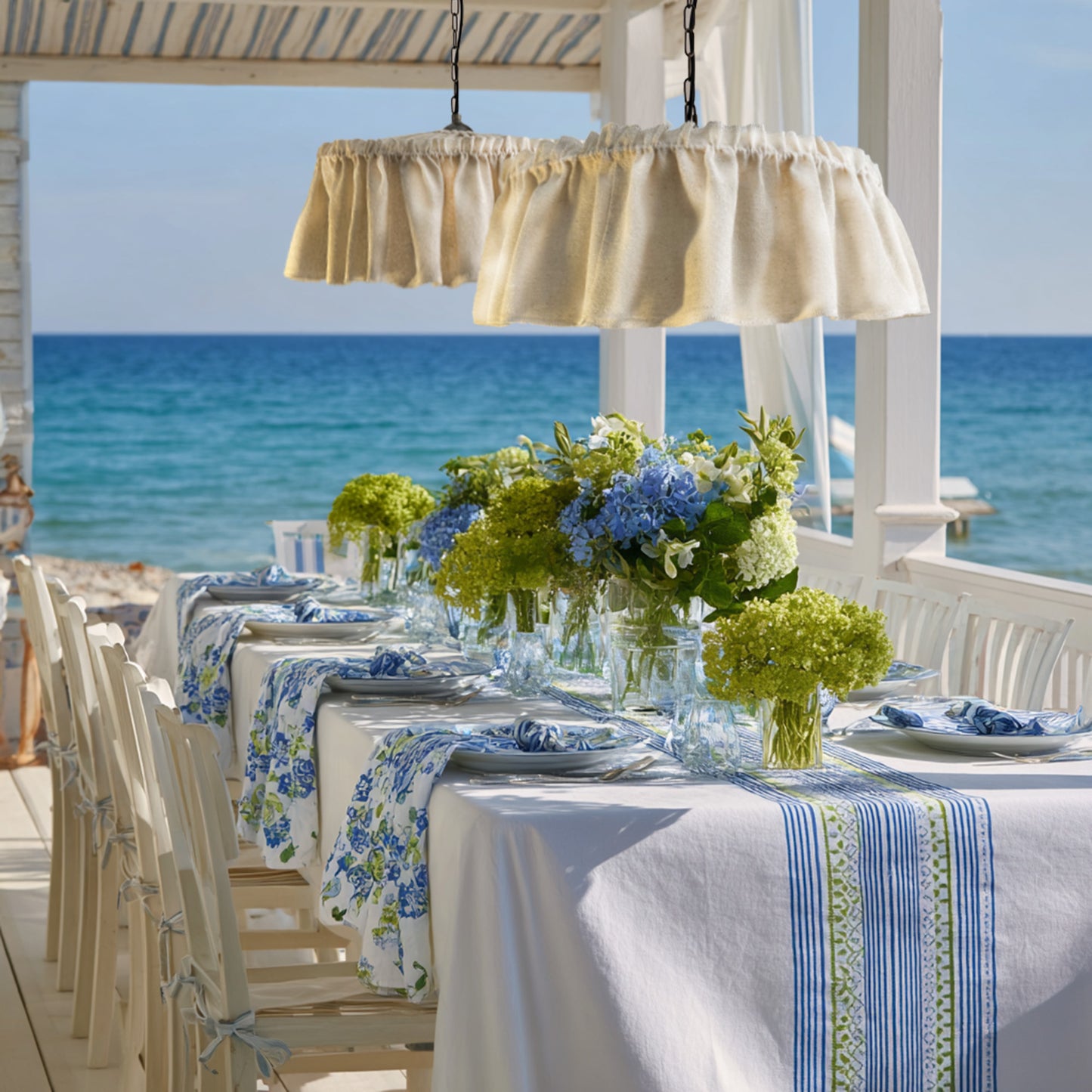 Ruffled raw silk lampshades hung over the dining table in beach cottage set with floral arrangements and striped tablecloth by the ocean.