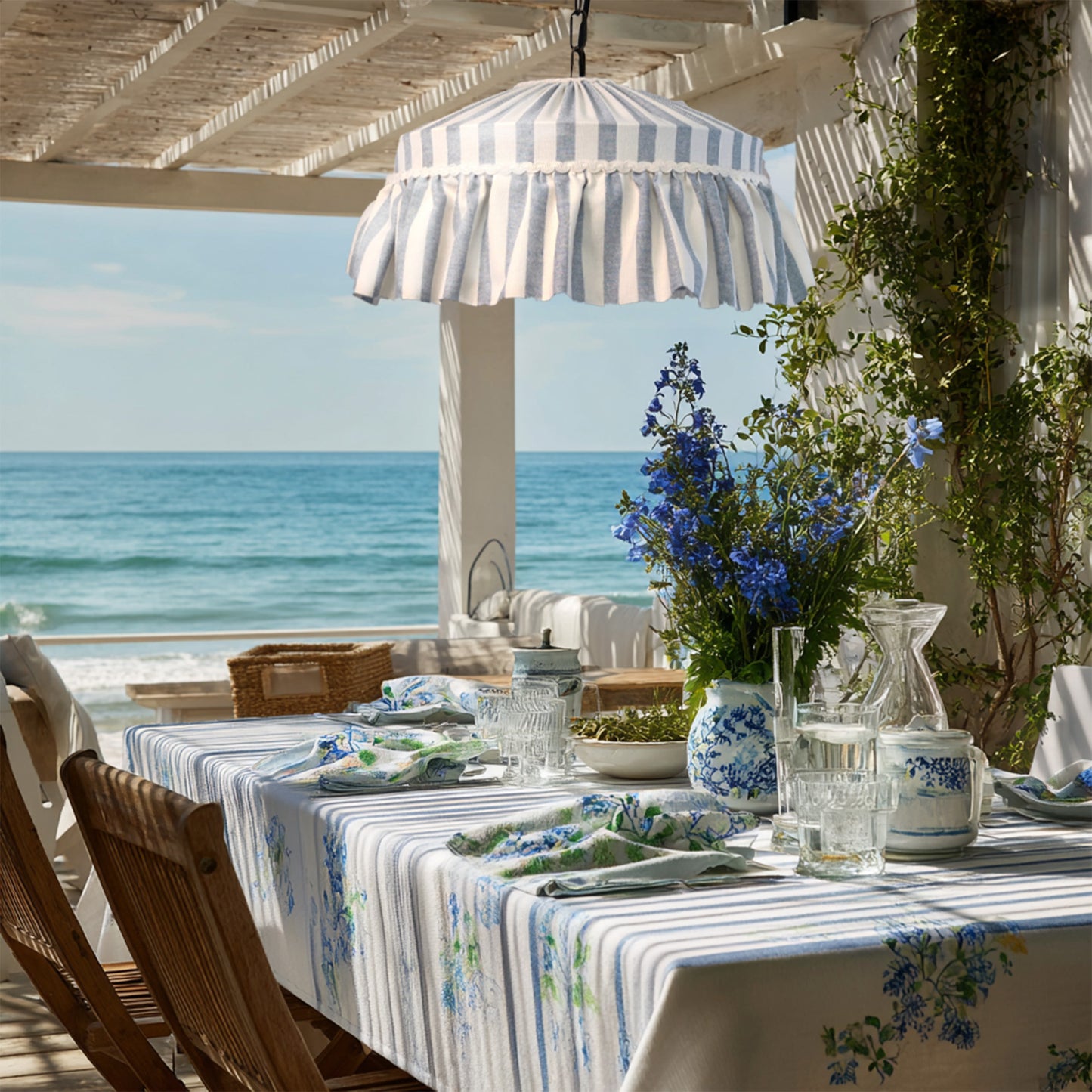 ruffle lampshade hung over outdoor dining table set with striped tablecloth and chairs, overlooking the ocean in beach cottage