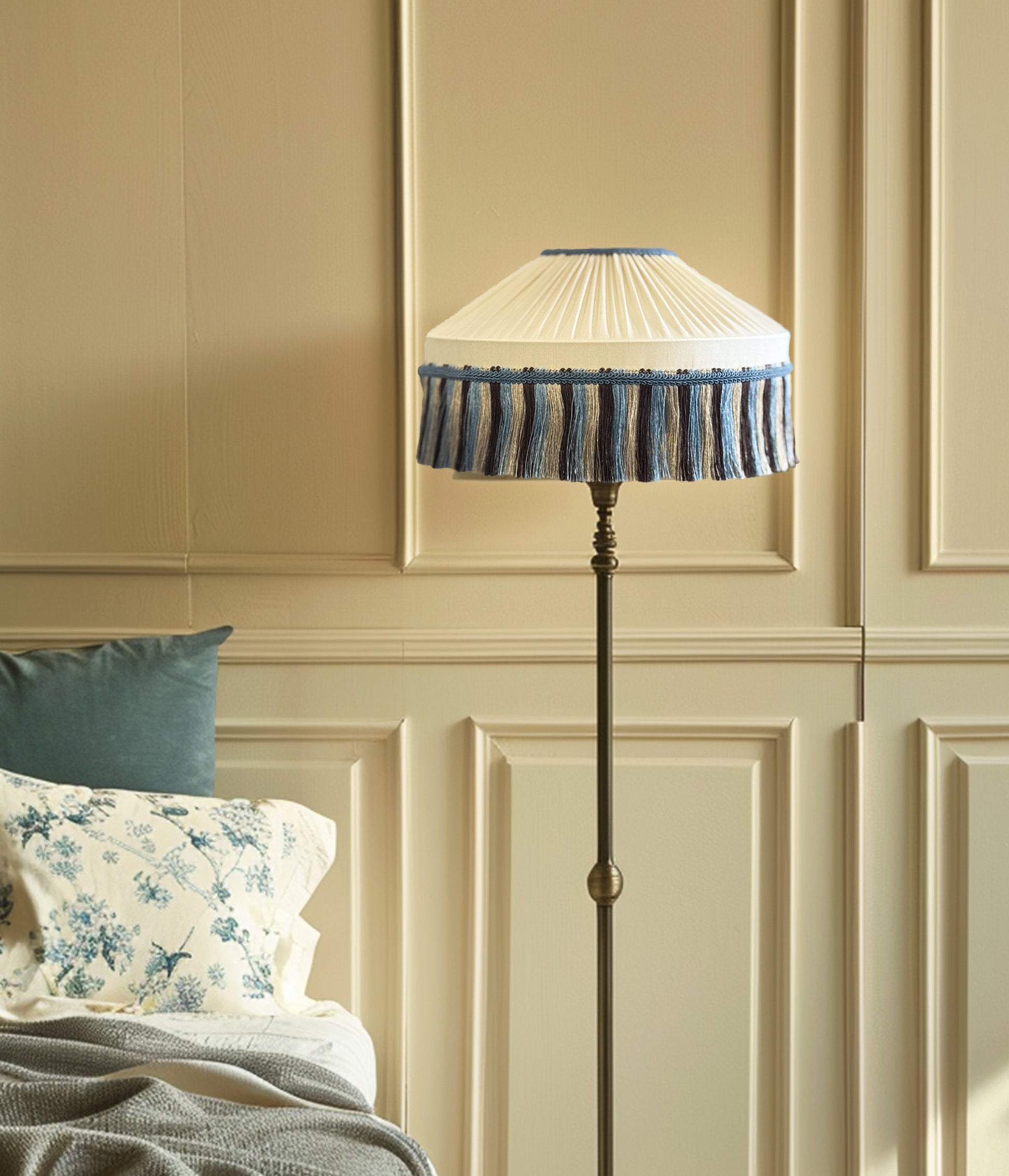 Floor lamp with decorative creamy with fringe lampshade in a room with beige paneled walls and floral pillows.